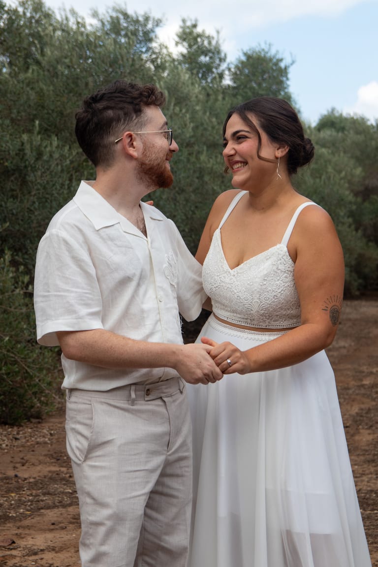 הכלה והחתן אוחזים ידיים ומביטים זה בזה בין עצי זית