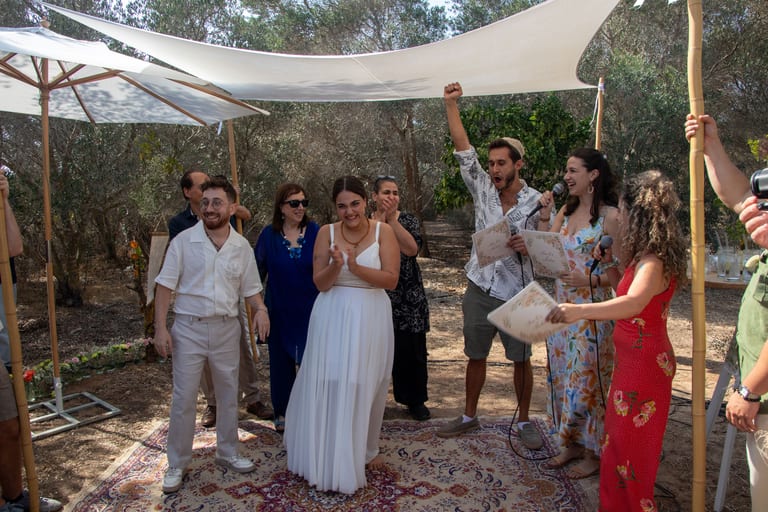 הכלה והחתן עומדים עם אורחים מתחת לסוכך בחורשה
